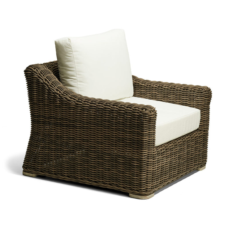 Sillón de mimbre marrón con cojines gruesos de color beige claro en el asiento y respaldo. El mueble presenta un diseño robusto y curvo, ideal para espacios exteriores o terrazas.