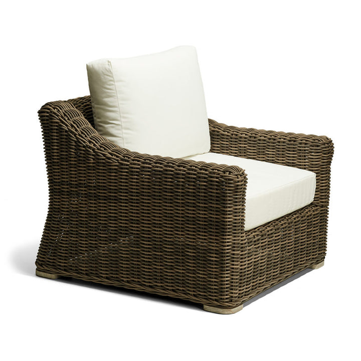 Sillón de mimbre marrón con cojines gruesos de color beige claro en el asiento y respaldo. El mueble presenta un diseño robusto y curvo, ideal para espacios exteriores o terrazas.