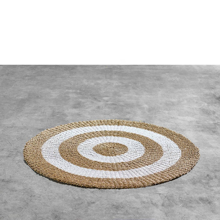 Alfombra redonda de fibra natural tejida a mano con un diseño de anillos concéntricos alternando entre marrón claro natural y blanco. El patrón simétrico proporciona un estilo decorativo moderno y orgánico.