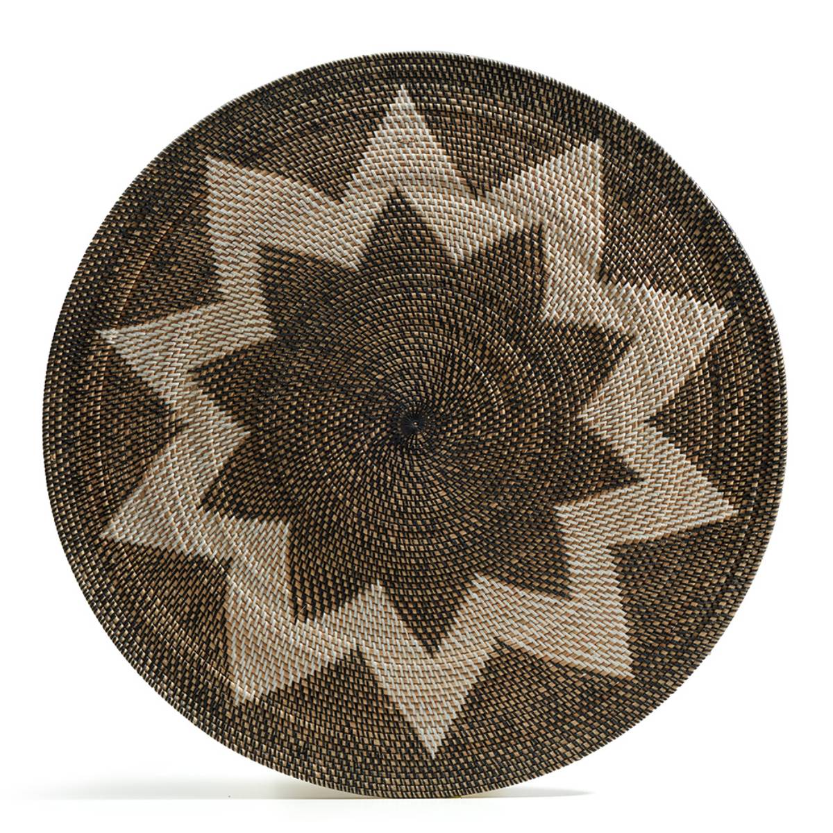 Canasta circular tejida a mano con patrón geométrico en forma de estrella de puntas pronunciadas, compuesta por fibras en tonos marrón, beige y negro.