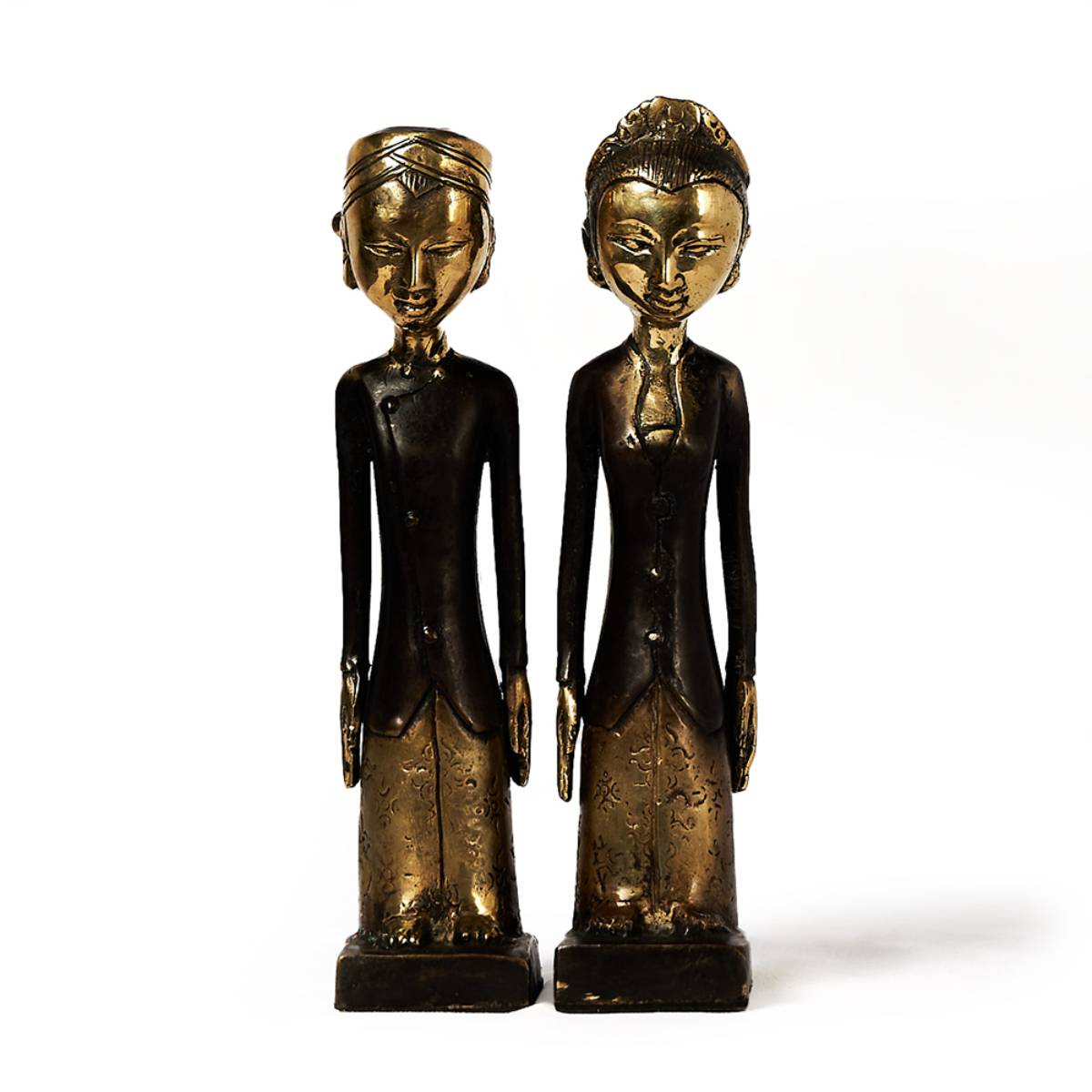 Dos figuras escultóricas alargadas y estilizadas, una con turbante y la otra con peinado recogido, ambas con trajes largos detallados, en tonos de bronce oscurecido y dorado. Las esculturas tienen detalles grabados en la ropa y expresión serena en los rostros.