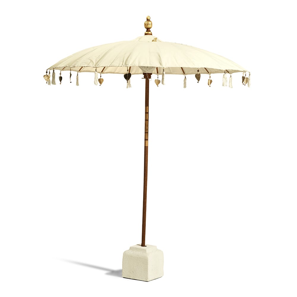 Paraguas decorativo de estilo balinés con tela color crema, detalles colgantes en forma de borlas y corazones, base cuadrada de cemento claro y mástil de madera terminado en un adorno dorado en la parte superior.