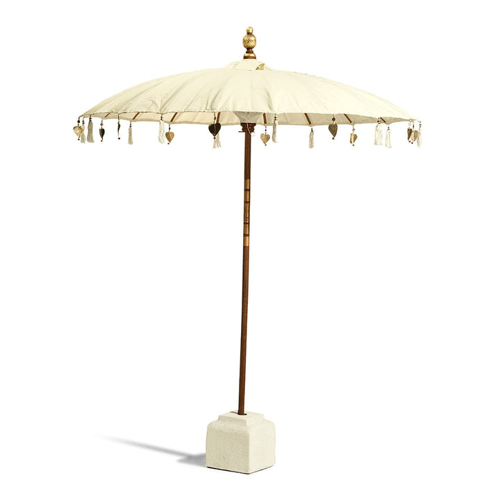 Paraguas decorativo de estilo balinés con tela color crema, detalles colgantes en forma de borlas y corazones, base cuadrada de cemento claro y mástil de madera terminado en un adorno dorado en la parte superior.