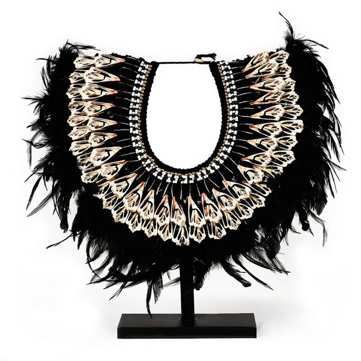 Collar decorativo en forma de media luna con múltiples plumas negras y detalles ornamentales en tonos claros y rosados, dispuesto en un soporte de exhibición. Los elementos decorativos incluyen patrones simétricos y tiras de cuentas en blanco y negro.