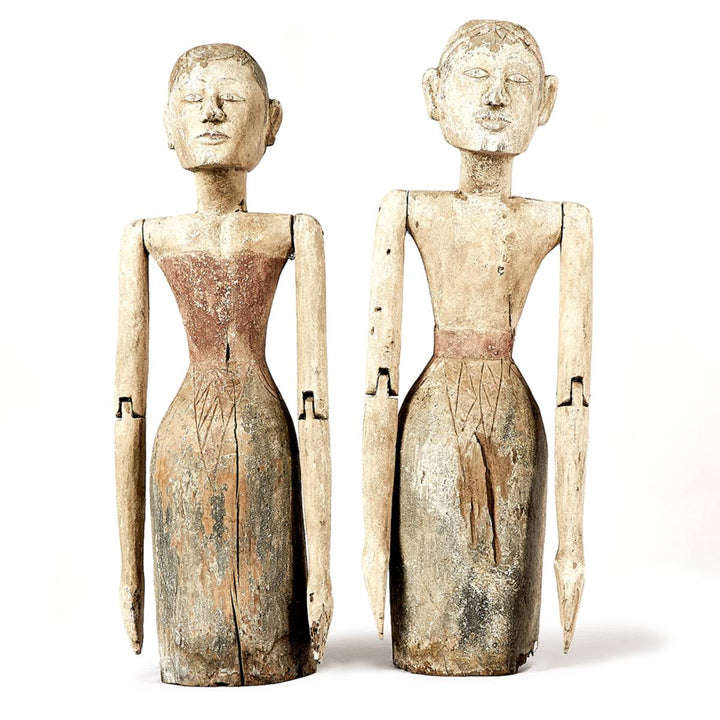 Dos figuras talladas en madera con forma humana, de aspecto estilizado y acabados rústicos, pintadas en tonos tierra. Ambas tienen brazos articulados y detalles grabados en la parte inferior del torso que sugieren vestimenta.
