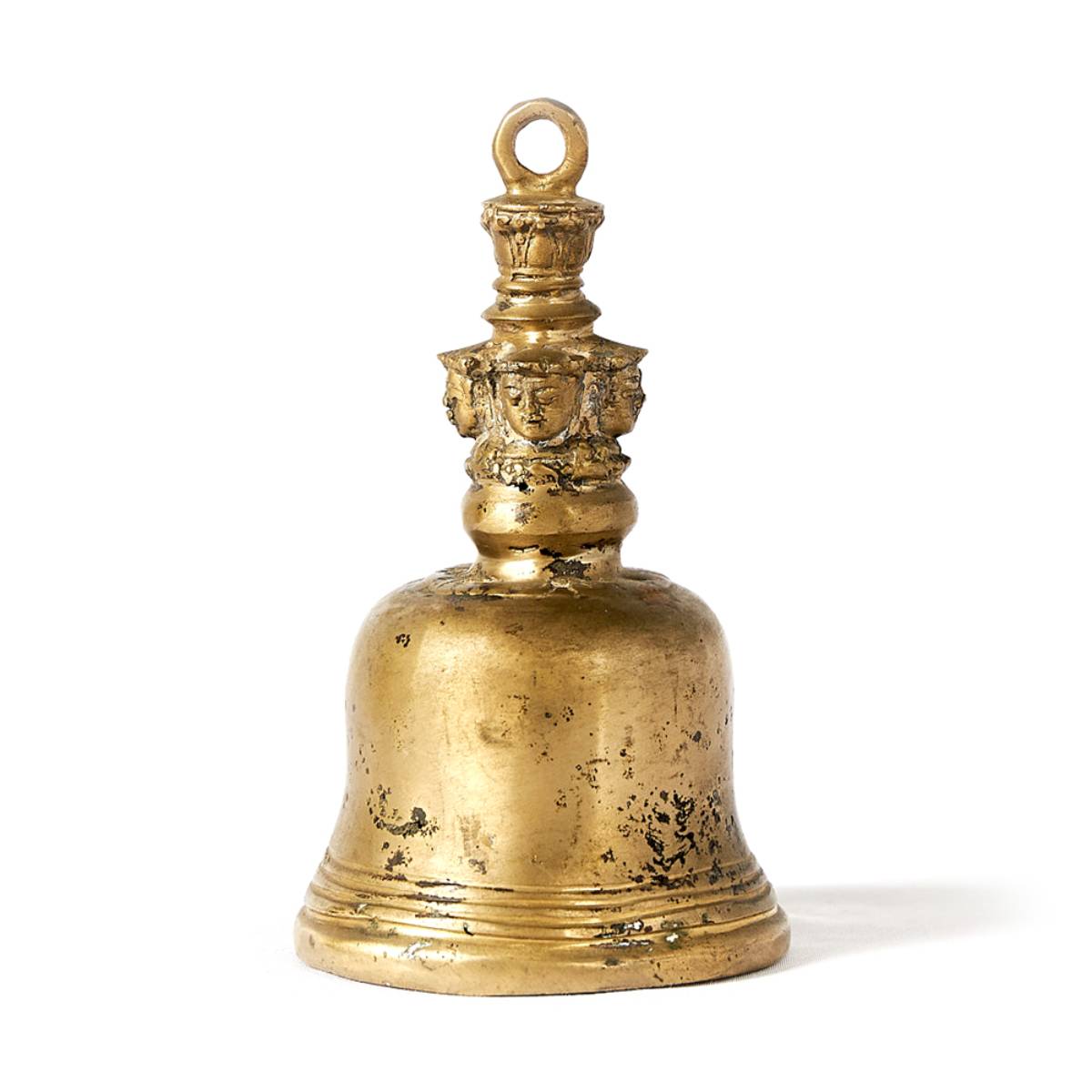 Campana de bronce dorado con acabado envejecido. El mango presenta un adorno con figuras talladas en relieve que representan rostros humanos estilizados.