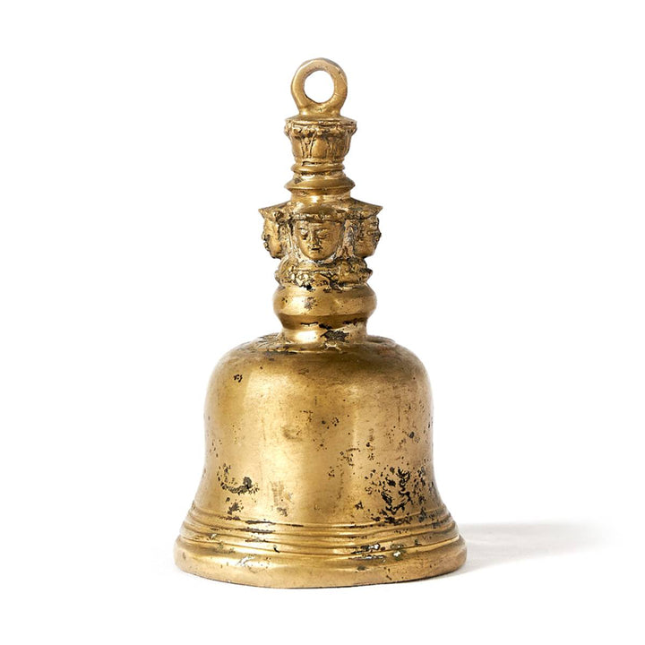 Campana de bronce dorado con acabado envejecido. El mango presenta un adorno con figuras talladas en relieve que representan rostros humanos estilizados.