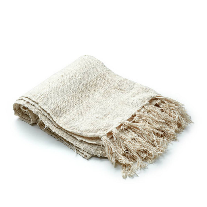 Una manta tejida de color beige claro con textura rústica y flecos en los extremos, doblada ordenadamente. No hay texto visible en el producto.