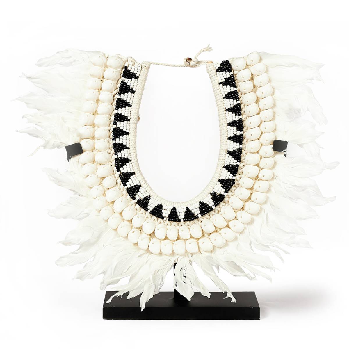 Collar decorativo con múltiples hileras de conchas blancas, cuentas negras y blancas dispuestas en un patrón geométrico, y una base de plumas blancas. Montado en un soporte negro, se utiliza como pieza ornamental.