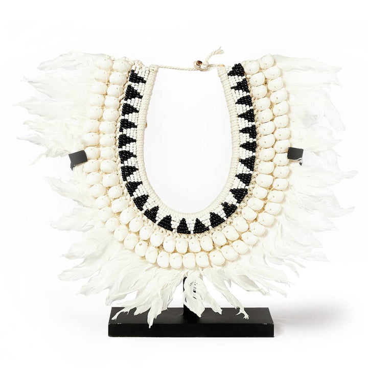 Collar decorativo con múltiples hileras de conchas blancas, cuentas negras y blancas dispuestas en un patrón geométrico, y una base de plumas blancas. Montado en un soporte negro, se utiliza como pieza ornamental.