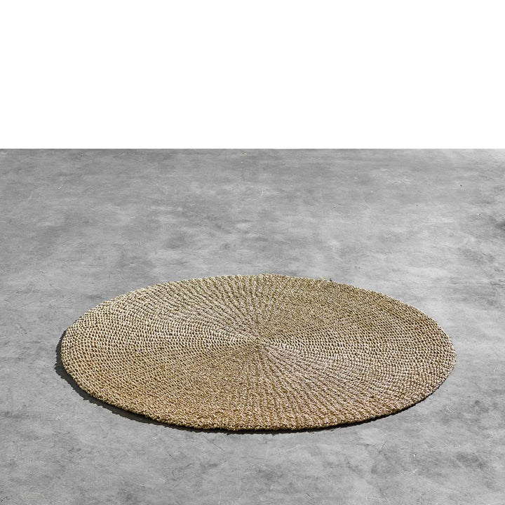 Alfombra redonda tejida de fibras naturales colocada sobre un piso de concreto gris; el diseño muestra un patrón radial desde el centro hacia los bordes.