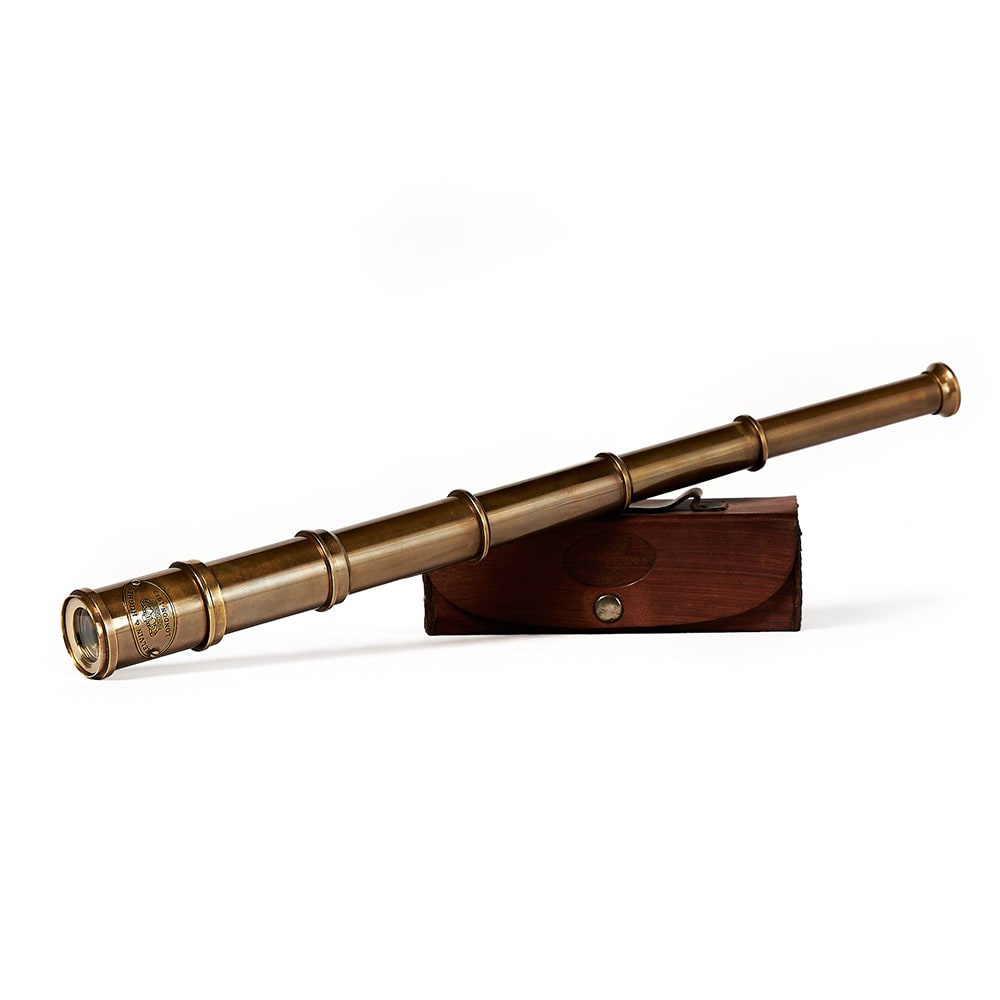 Un telescopio náutico extensible de bronce con un acabado envejecido, acompañado de una caja de madera oscura con cierre metálico. En uno de los extremos del telescopio se puede leer una inscripción grabada con la marca y detalles decorativos.