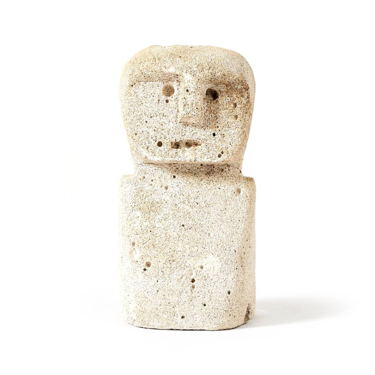 Figura tallada de piedra pómez con forma humanoide, de cuerpo rectangular y rostro simplificado con ojos, nariz y boca. La superficie es porosa y tiene un acabado rústico.