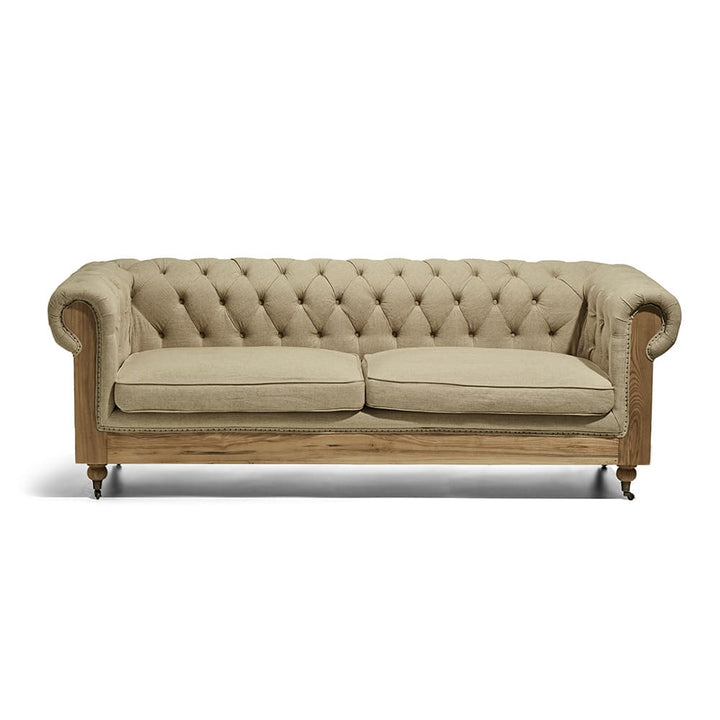 Sofá estilo Chesterfield de tres cuerpos con acolchado capitoné y tapizado en tela beige. Tiene una estructura de madera visible y ruedas pequeñas en las patas delanteras.