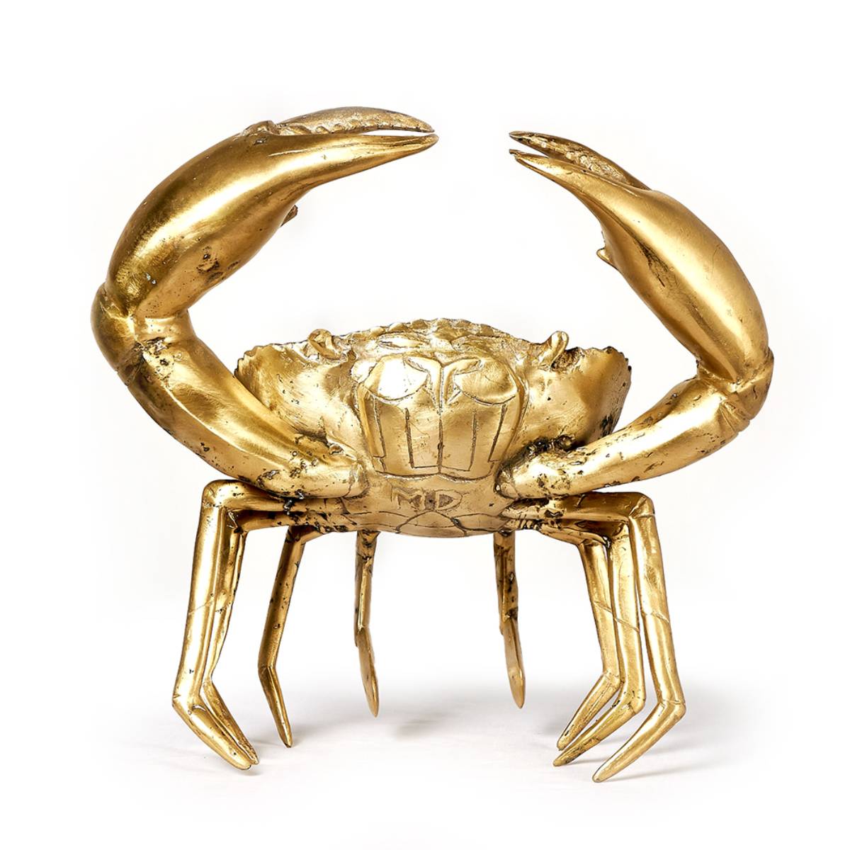Escultura de un cangrejo metálico dorado con garras levantadas, detalles realistas y las letras "MD" grabadas en el centro del caparazón. La pieza presenta un acabado brillante con zonas de desgaste.