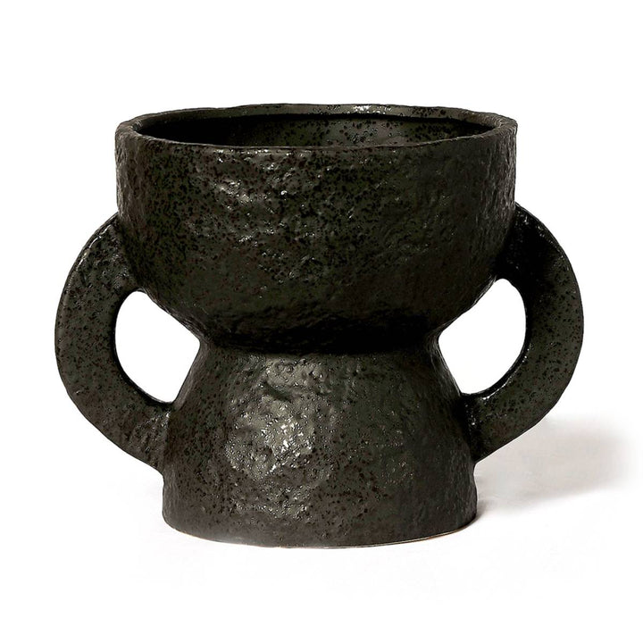 Taza de cerámica con superficie rugosa y acabado en negro oscuro, presenta un diseño simétrico con dos asas circulares más grandes de lo habitual. Su forma se asemeja a un reloj de arena con un borde superior irregular.