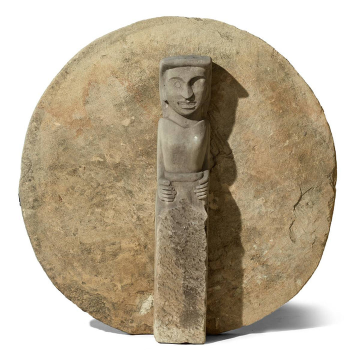 Escultura de piedra que representa una figura humana estilizada con una expresión facial marcada y brazos detallados, colocada frente a un gran disco de piedra.