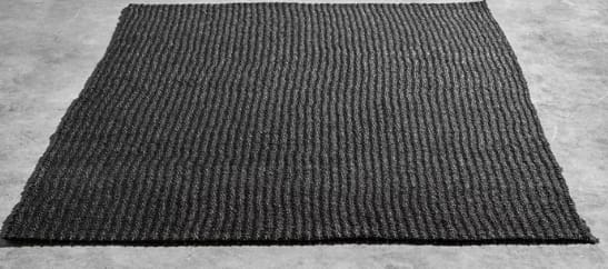 Alfombra cuadrada de textura gruesa con patrón de líneas verticales en tonos oscuros, colocada sobre un suelo gris claro. No presenta detalles escritos visibles.