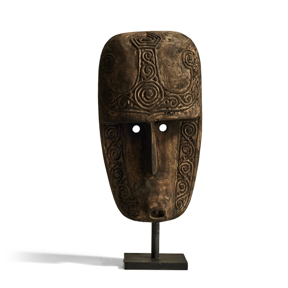 Máscara tallada de madera con intrincados patrones en espiral, motivos simétricos y dos orificios redondos para los ojos; se presenta montada sobre un soporte negro.