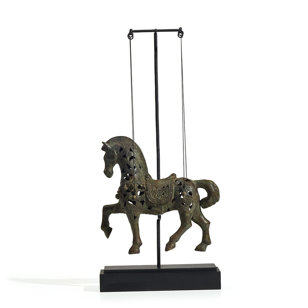 Escultura decorativa de un caballo en posición de marcha, elaborada en metal con acabados en bronce envejecido y patrones calados en el cuerpo. Está suspendida por dos delgados cables de un soporte vertical con base rectangular negra.