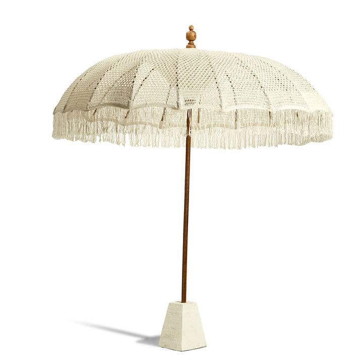 Sombrilla de pie con estructura de madera y una cubierta de tejido tipo macramé en color beige claro, con flecos decorativos colgando del borde. La base parece ser de cemento en forma piramidal truncada.