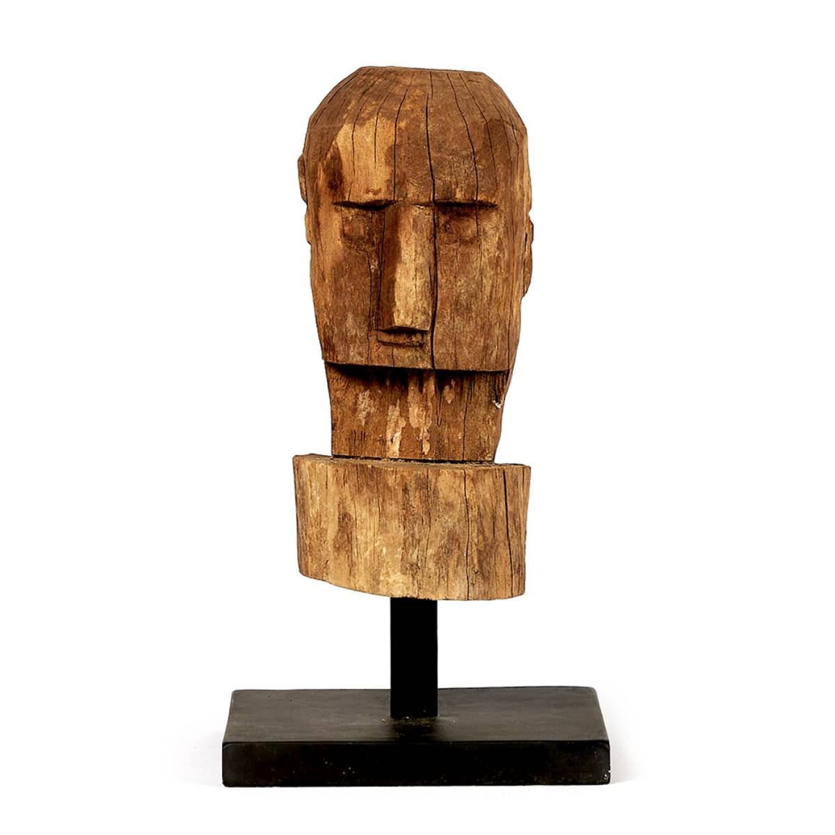 Escultura de madera con forma de cabeza humana estilizada y geométrica, colocada sobre una base negra rectangular. La figura presenta un acabado rústico con grietas visibles y vetas naturales de la madera.