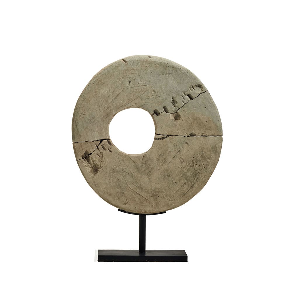 Disco de piedra circular con un agujero en el centro, montado sobre un soporte metálico negro. La superficie tiene grietas visibles y textura rugosa, lo que sugiere un estilo rústico o antiguo.