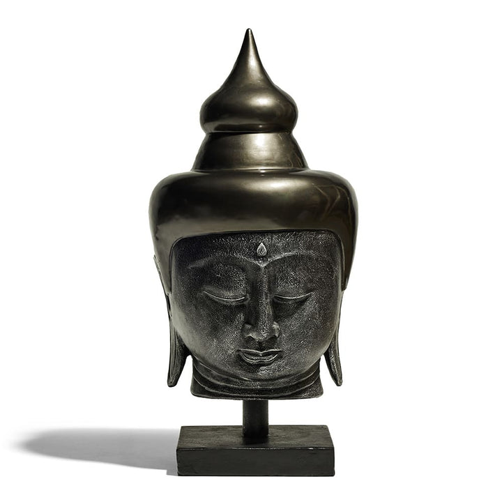 Escultura de una cabeza de Buda con acabado metálico oscilando entre tonos oscuros y brillantes, montada sobre una base rectangular. El rostro muestra expresión serena con ojos cerrados y un detalle en la frente.