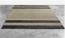 Alfombra rectangular con patrón de rayas horizontales en tonos beige y gris oscuro, tejida con una textura natural y aspecto rústico.