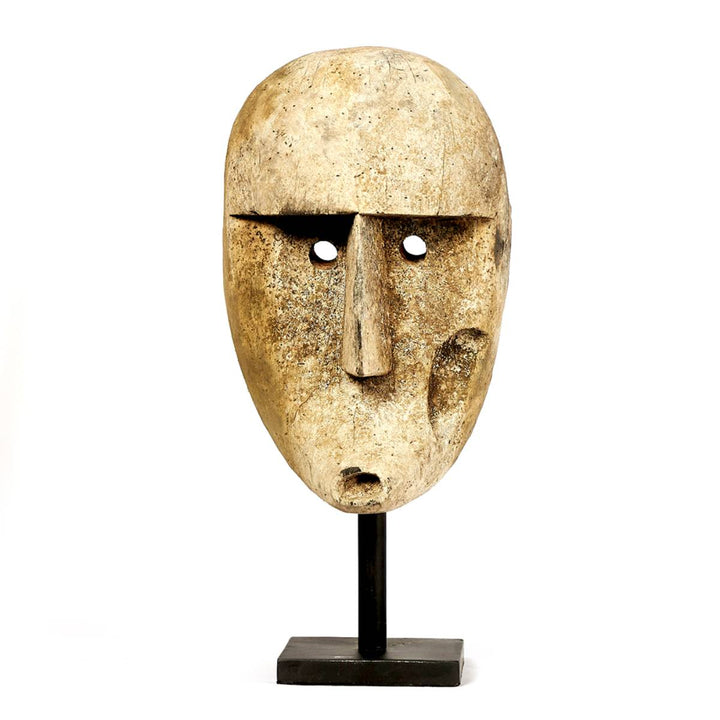 Máscara tribal tallada en madera de forma ovalada, con ojos circulares perforados, nariz y boca prominentes, montada sobre una base negra rectangular. Su superficie muestra un acabado envejecido con tonos terrosos y texturas rústicas.