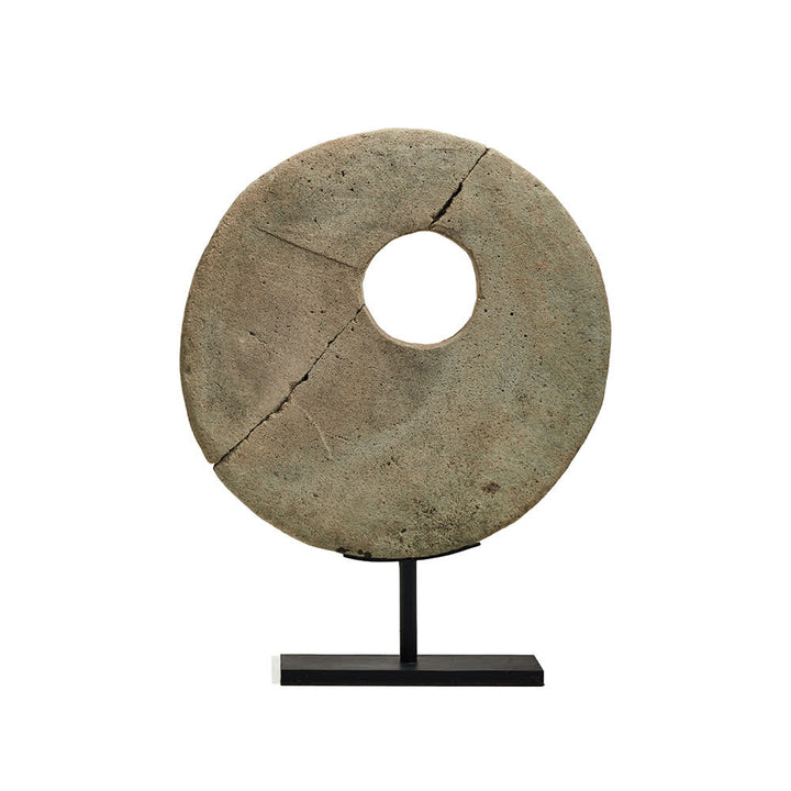 Objeto decorativo circular de piedra con un agujero en el centro, montado sobre una base metálica negra; la piedra muestra grietas visibles que indican desgaste y antigüedad.