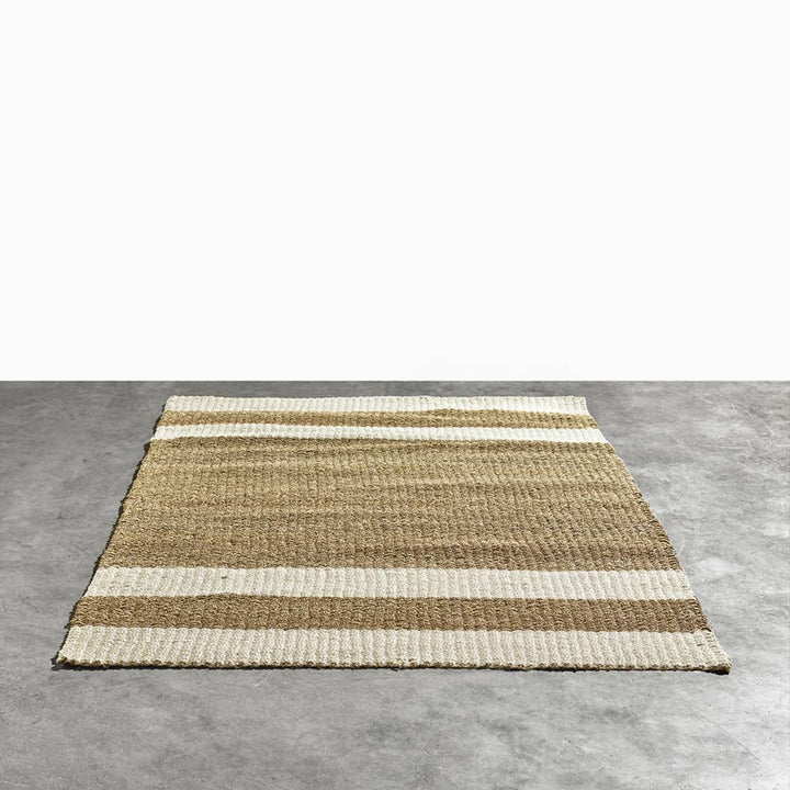 Alfombra rectangular tejida con bandas horizontales en tonos beige y marrón claro, colocada sobre un suelo gris de cemento pulido.