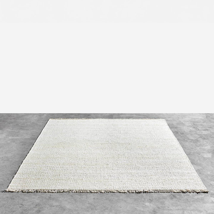 Alfombra rectangular de tejido fino color blanco roto con flecos cortos en los bordes más estrechos. Está colocada sobre un piso de cemento gris.