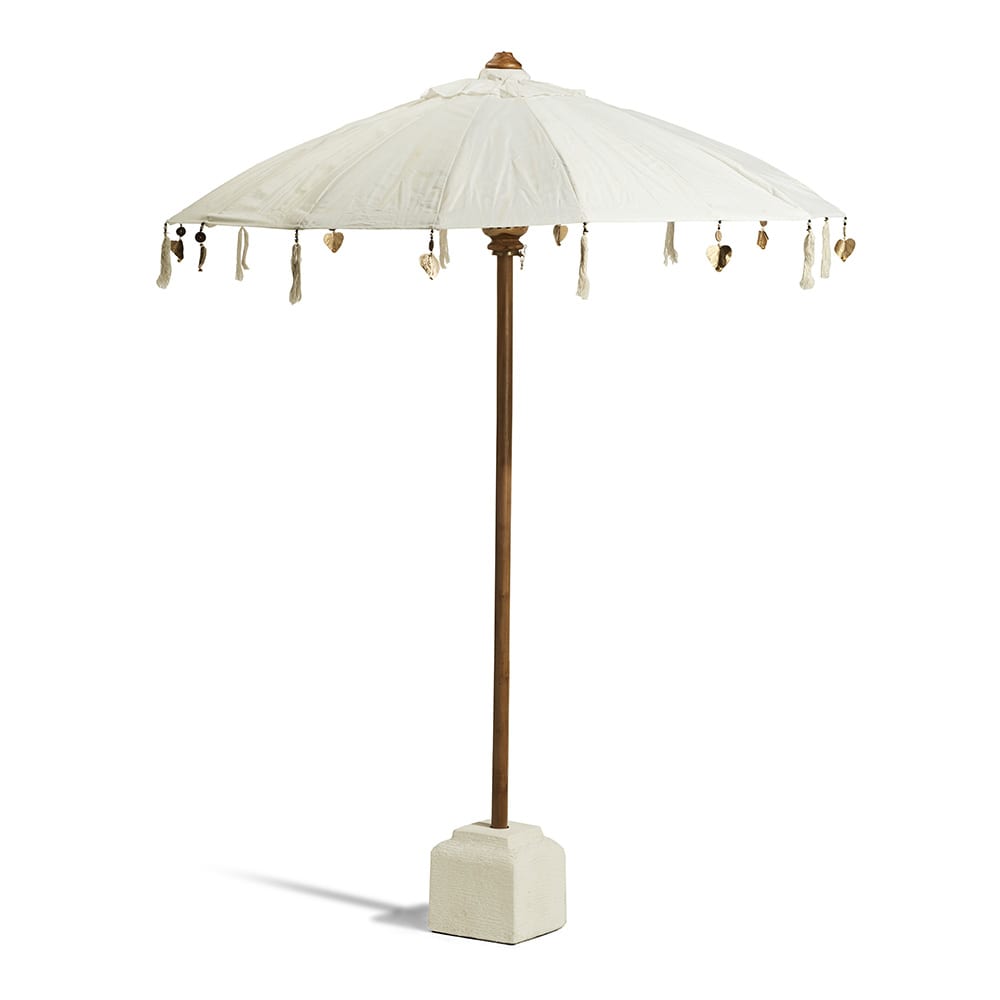 Sombrilla de estilo rústico con tela blanco hueso, soporte de madera y base cuadrada de cemento claro. Cuelgan adornos decorativos en los bordes de la tela, incluyendo borlas claras y pequeñas figuras doradas.