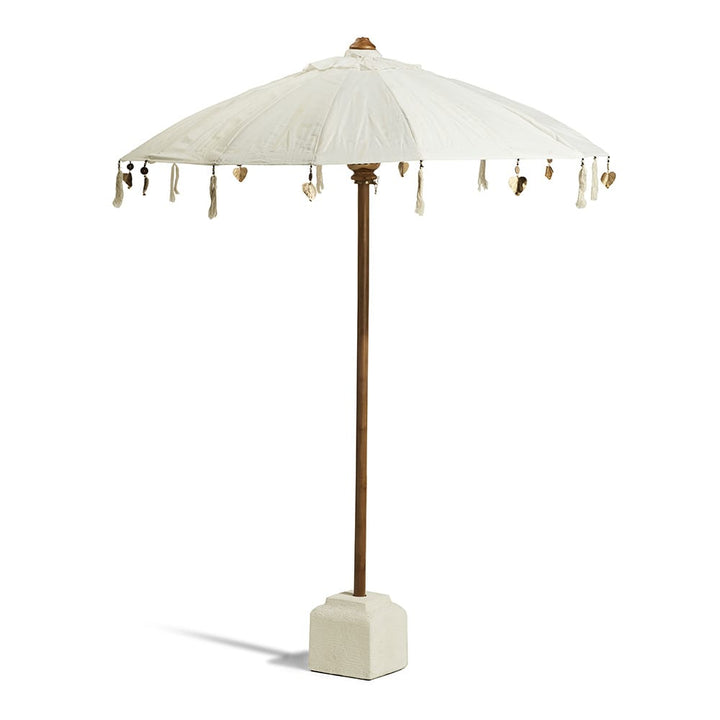 Sombrilla de estilo rústico con tela blanco hueso, soporte de madera y base cuadrada de cemento claro. Cuelgan adornos decorativos en los bordes de la tela, incluyendo borlas claras y pequeñas figuras doradas.