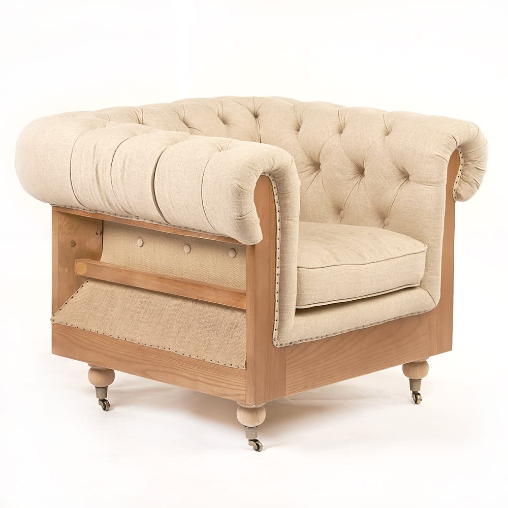Sillón acolchado estilo Chesterfield con tapizado en lino beige claro y estructura visible de madera clara, montado sobre ruedas pequeñas. El diseño presenta un respaldo y apoyabrazos abotonados y curvados, con acabado artesanal.